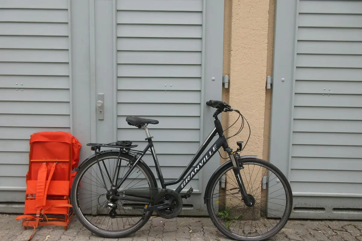Black hybrid bicycle parked in front of gray garage doors with a bright red bag leaning against the wall to the left.