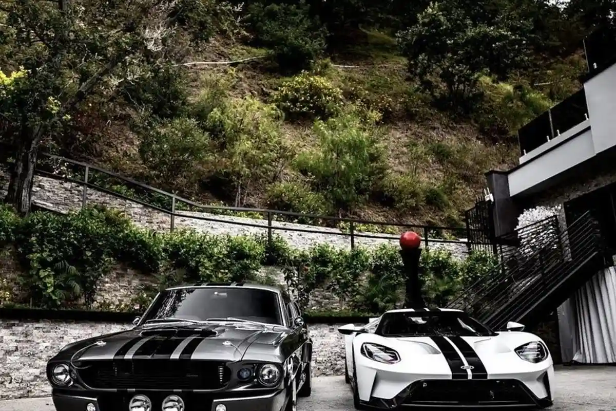 Two high-performance sports cars with black racing stripes parked in a modern garage courtyard, with a hillside and stairway in the background.