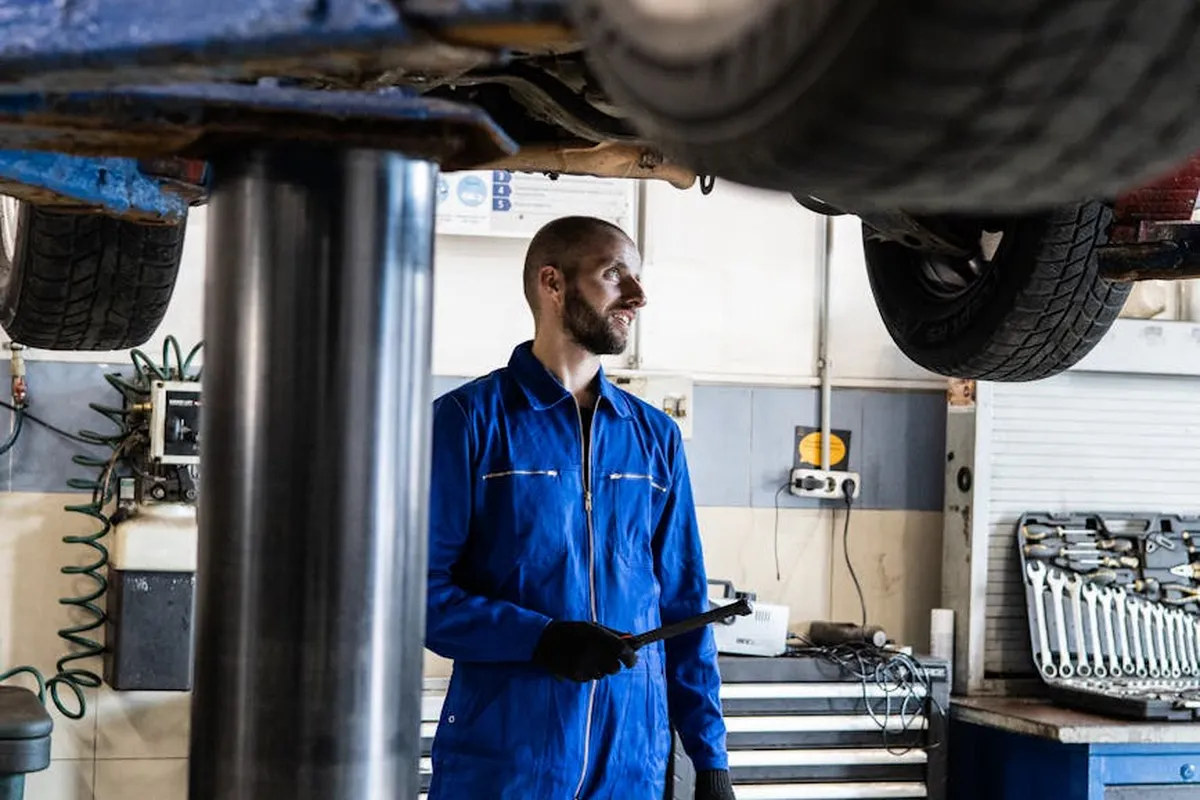 A mechanic in blue coveralls stands beside a lifted vehicle in a busy auto repair shop.
