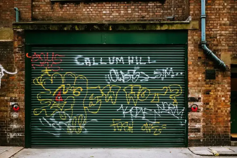 Green metal garage door with graffiti on a brick building, urban street scene.