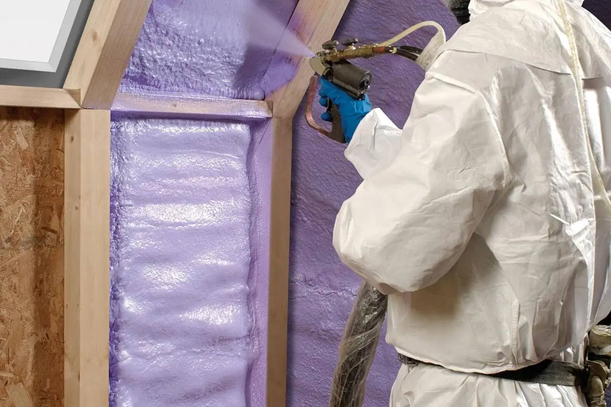 A worker in protective suit applying spray foam insulation to the interior garage wall near a doorway.