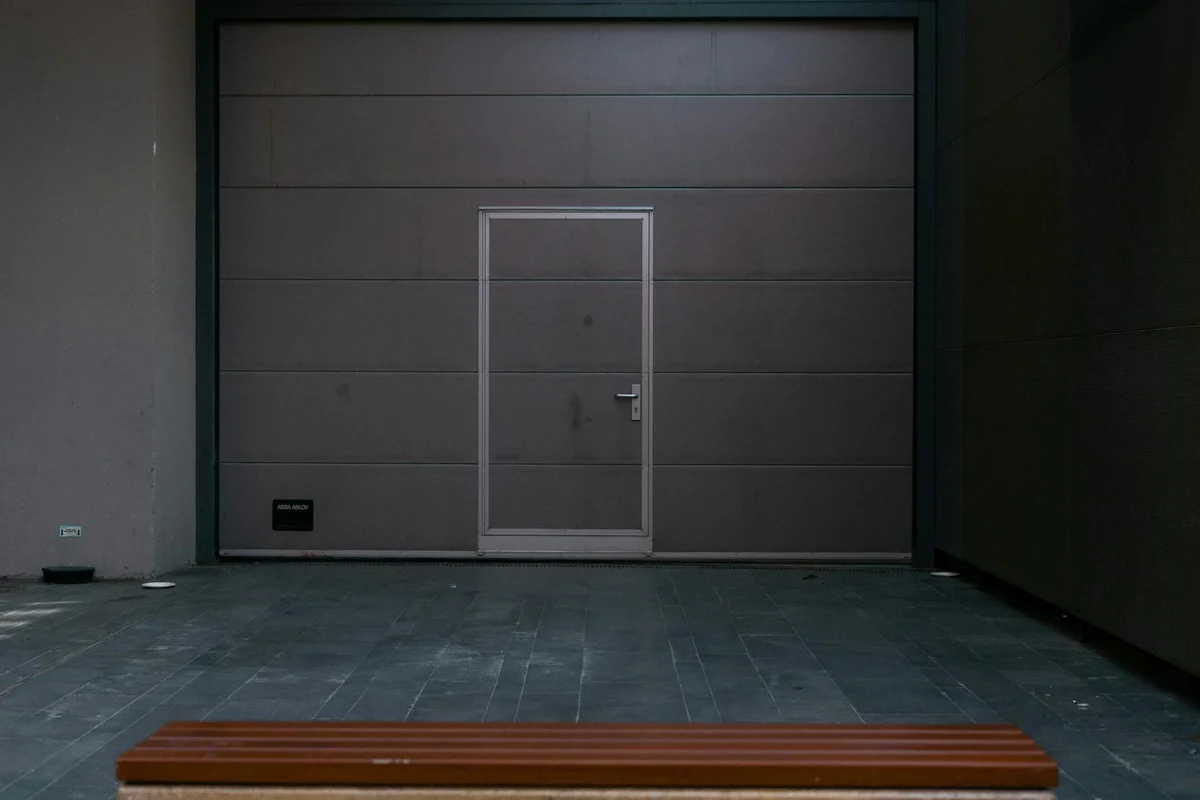 Indoor garage with a large gray sectional door and a small pedestrian door in the center; a wooden bench sits on the floor in front.