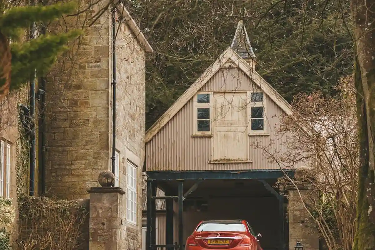 Residential garage with a red car parked inside under a steeply pitched wooden façade, surrounded by trees.