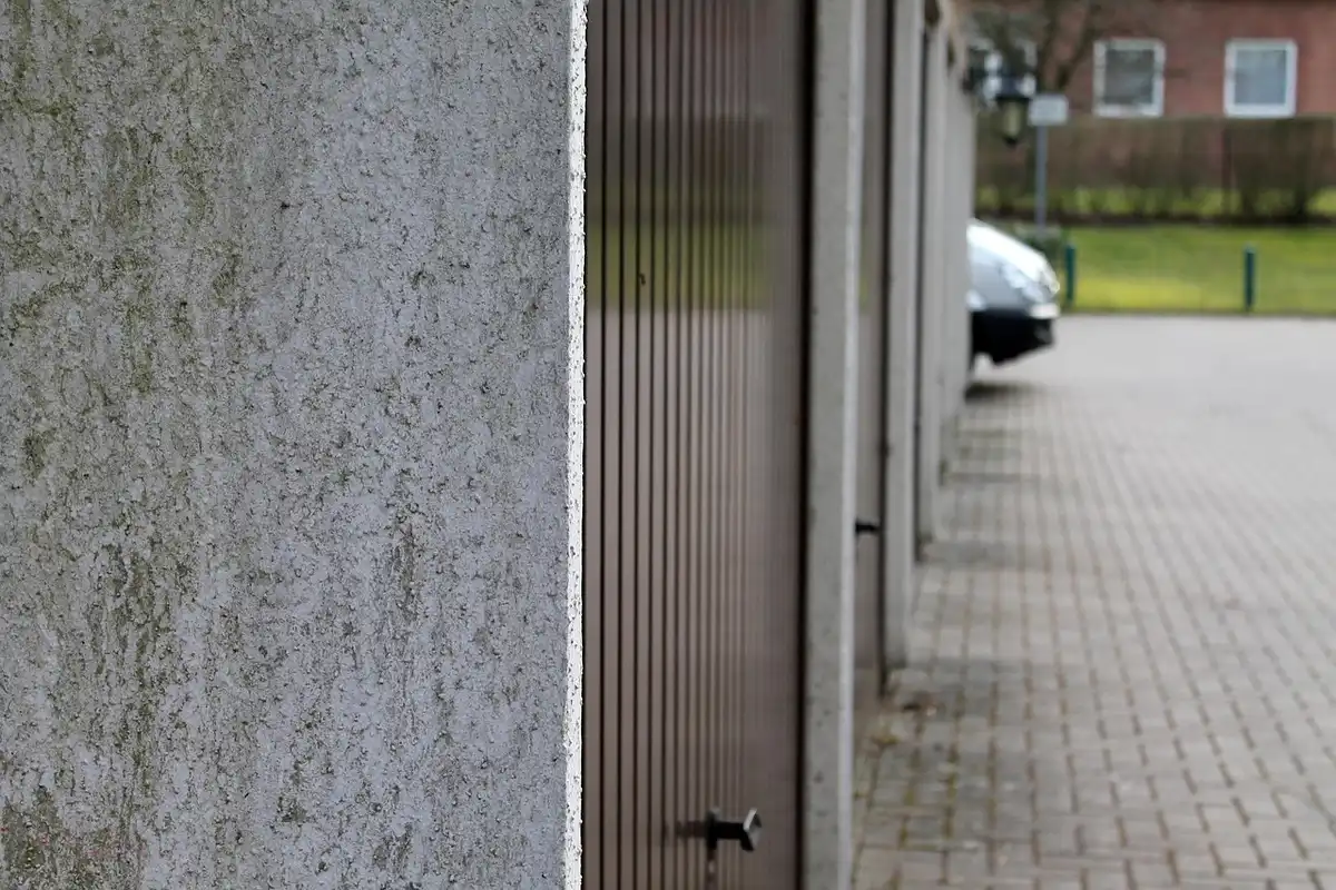 Garage door along a paved driveway with a concrete wall on the left and a car in the distance.