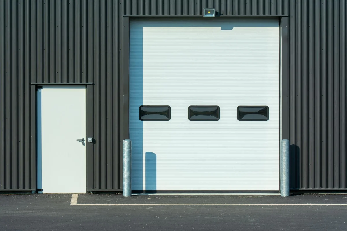 White sectional garage door with three small rectangular windows, a side entry door, and safety bollards against a dark metal building.