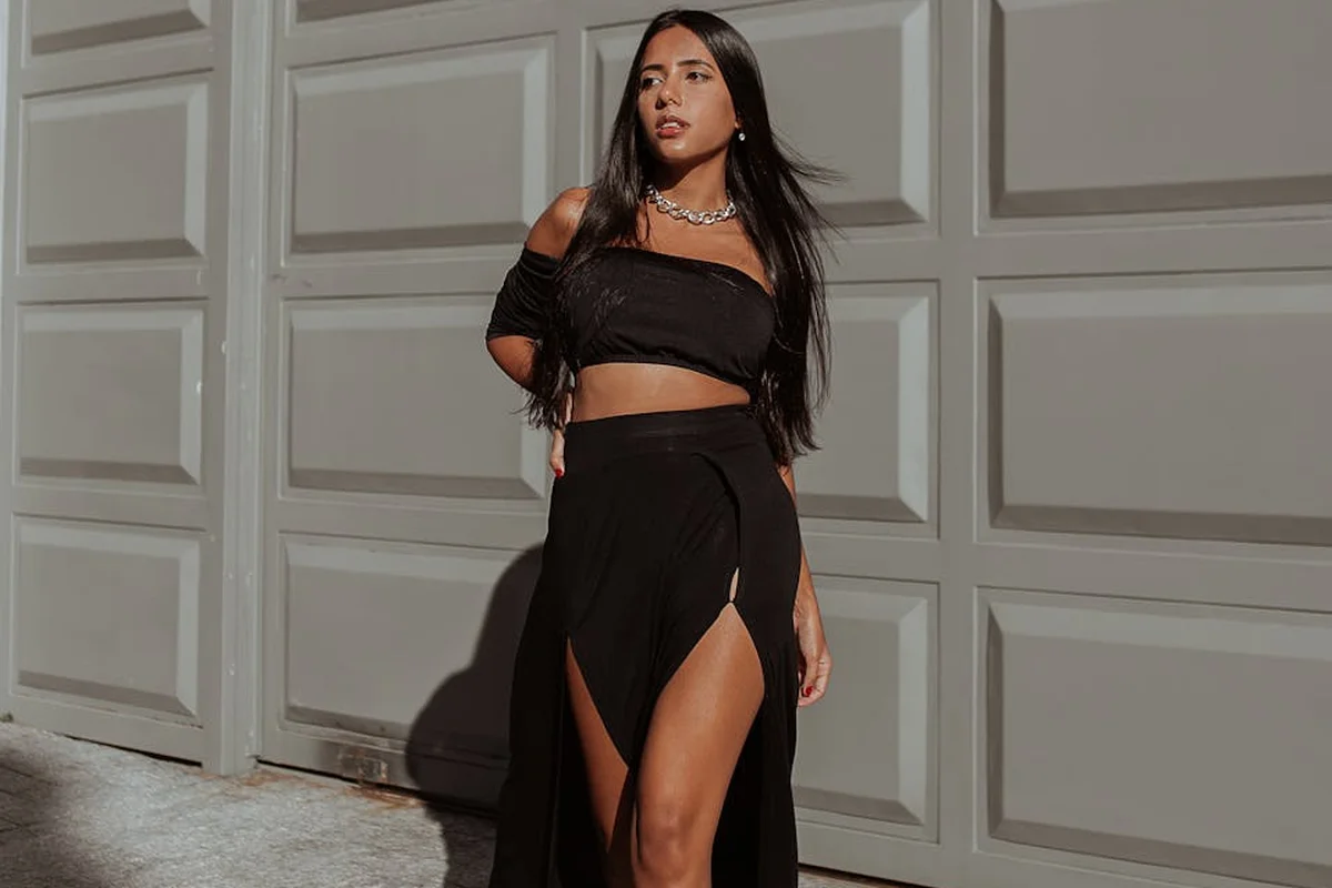 A person wearing a black crop top and a high-slit black skirt stands in front of a gray garage door.