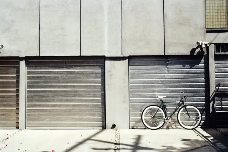 A row of grey metal garage doors; a bicycle leans against the right-hand door while sunlight casts shadows on the concrete driveway.