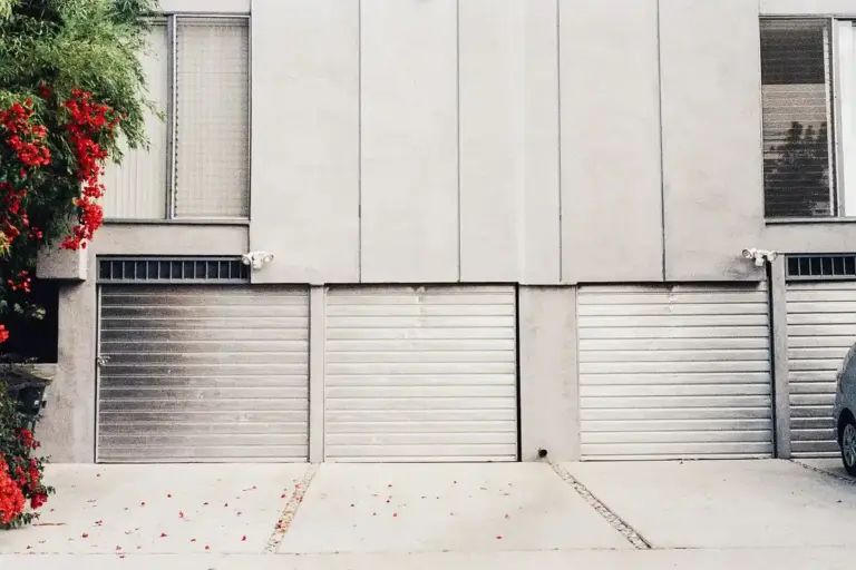 Row of gray sectional garage doors on a residential building with a driveway; a car is partially visible on the right and a flowering bush on the left.