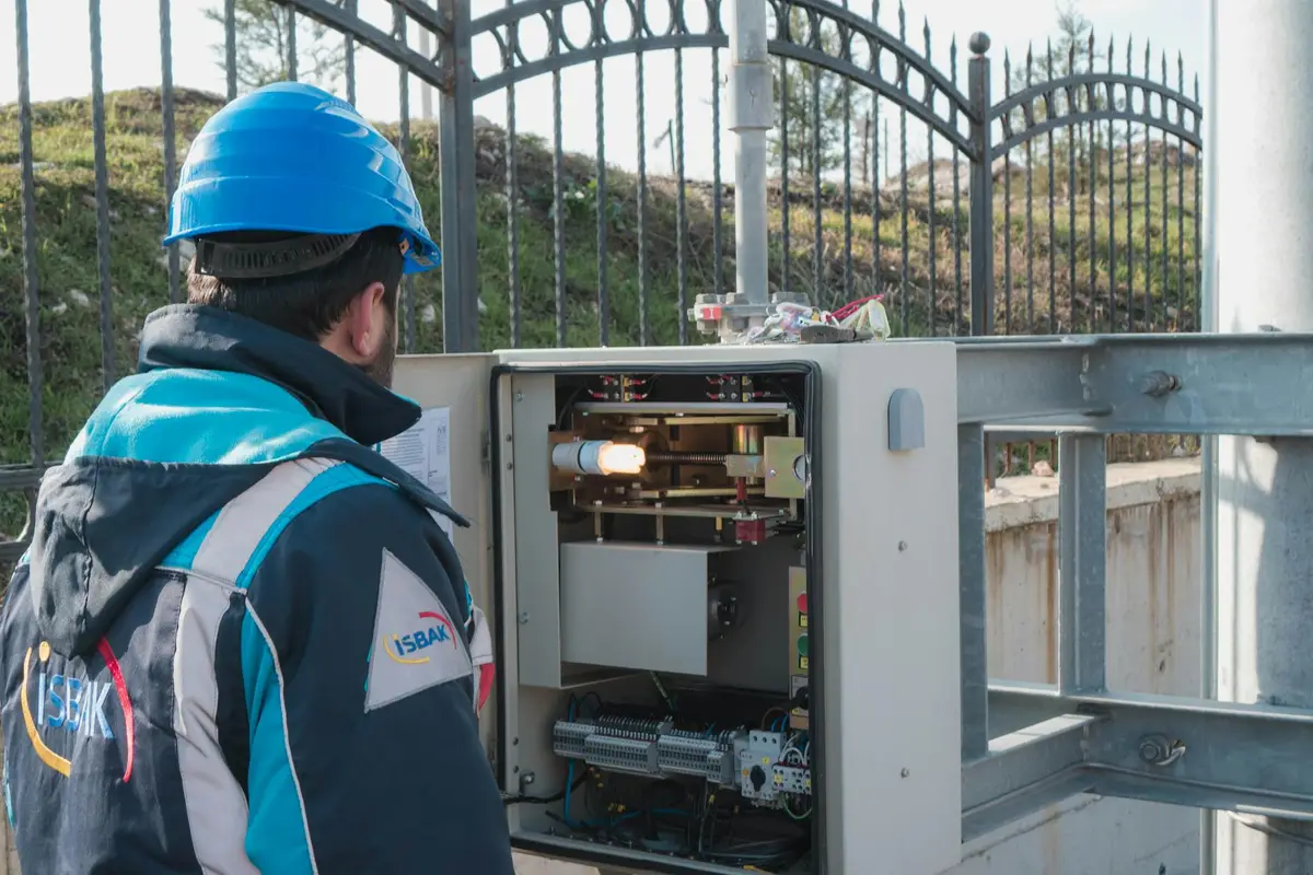 A technician wearing a blue hard hat and protective jacket inspects an outdoor electrical panel mounted on a metal frame near a fence.