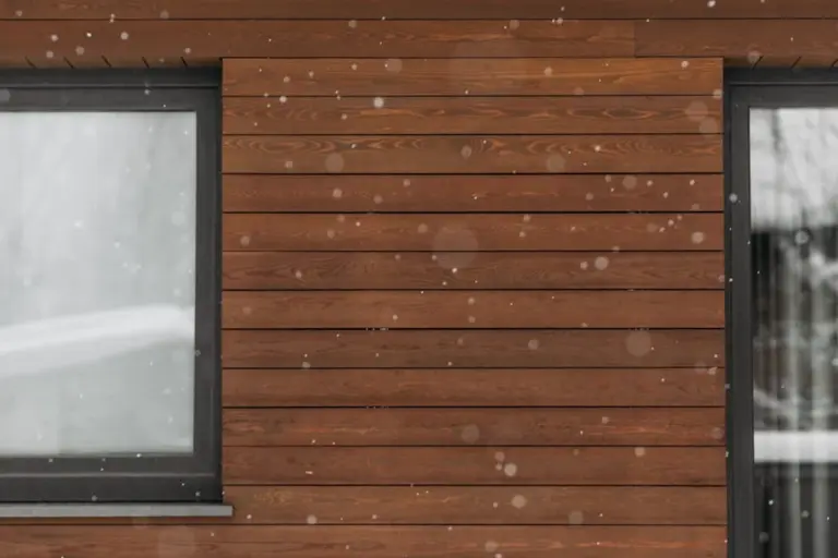 Brown wooden garage siding with a window on the left and a door on the right, with snowfall in winter.
