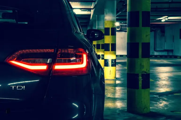 Dim underground parking garage with a dark blue car and red tail lights, yellow-and-black striped pillars.