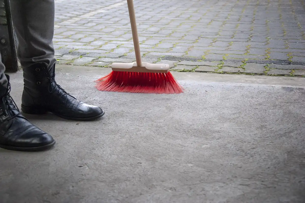 A red-bristled broom on a concrete garage floor, with a pair of black boots in the background