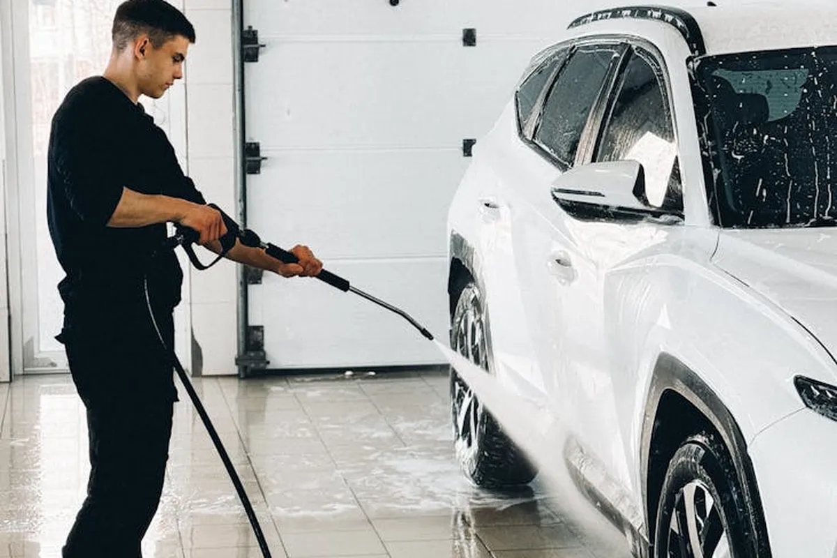 A person using a pressure washer to wash a white SUV inside a clean garage, with a wet floor.