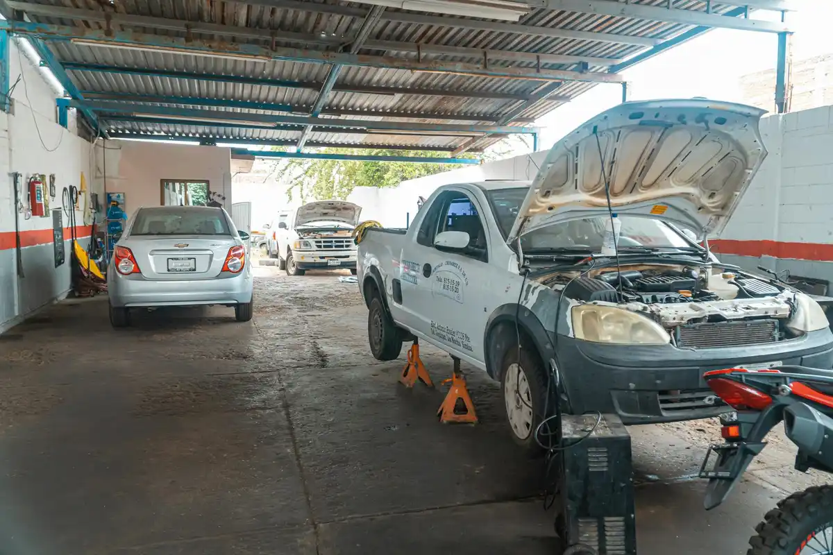 A vehicle with its hood open is supported by jack stands inside a covered garage, with another car parked in the background and a concrete floor showing wear.