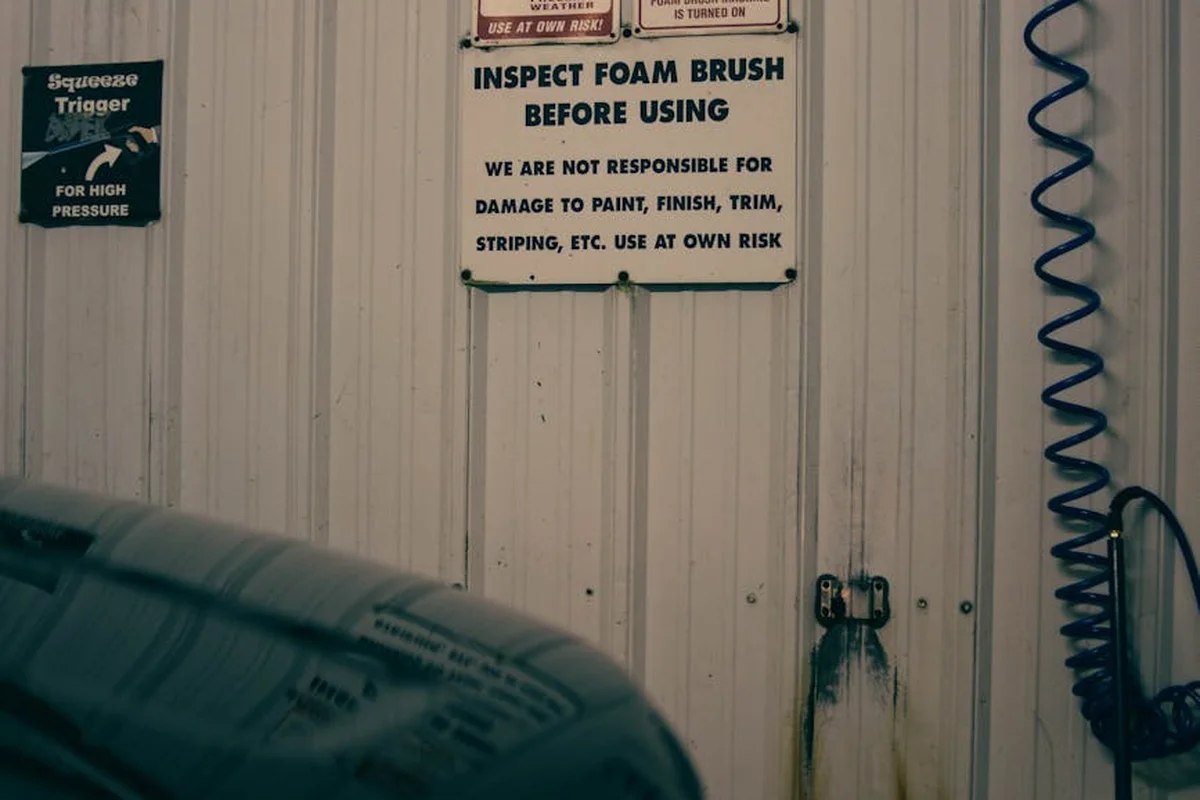 Garage workshop with safety signs on a metal wall, a coiled air hose on the right, and a car hood in the foreground, illustrating prep and safety considerations before coating the floor.