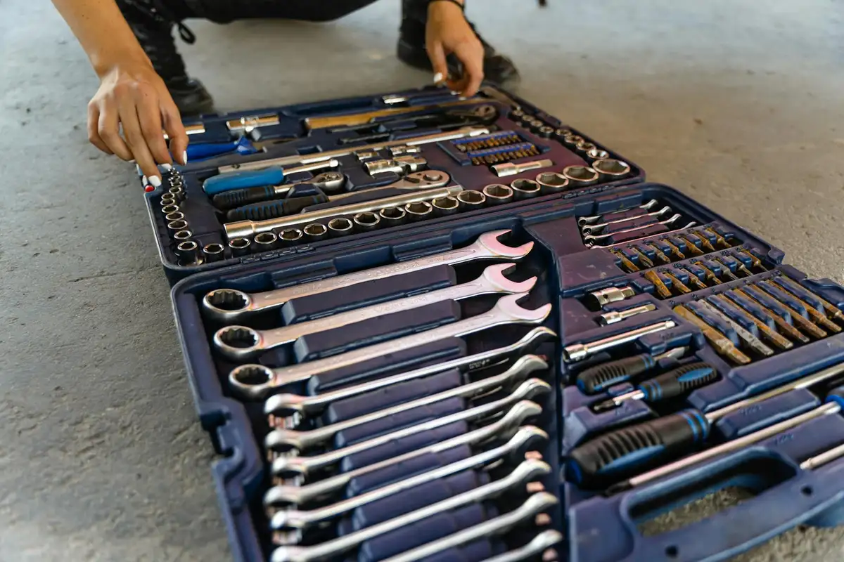 Close-up of a toolbox opened on a concrete garage floor, with various wrenches and sockets arranged neatly as a person reaches for a tool.