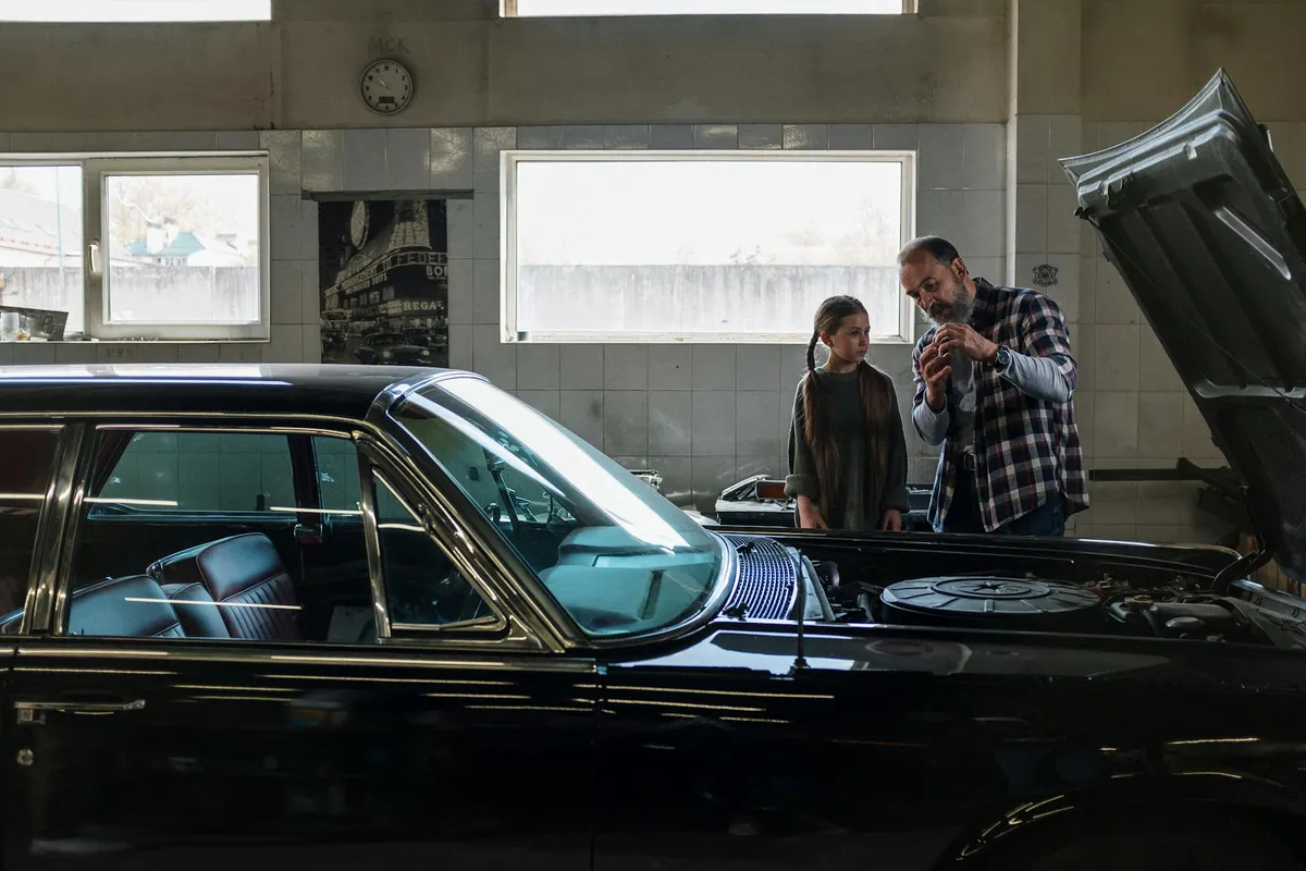 A man and a woman stand in a well-lit garage inspecting a car with its hood open.