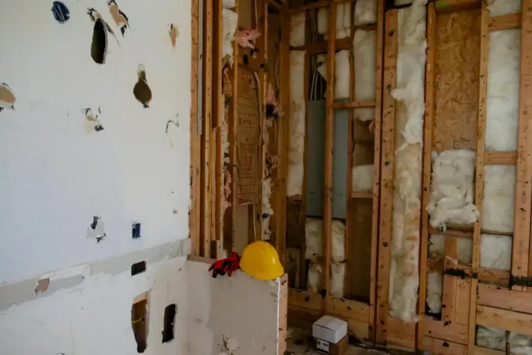 Open garage wall with exposed framing studs and fiberglass insulation; a yellow hard hat rests on a stud, indicating ongoing insulation work.