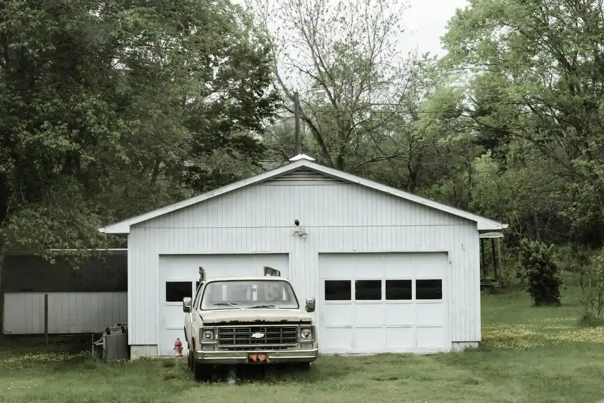 White two-car garage with a vintage pickup truck parked on a grassy front yard, surrounded by trees.