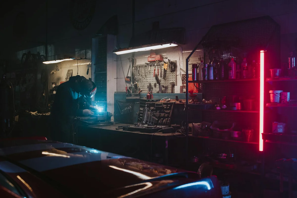 Dimly lit garage with a person near a car, illuminated by blue task lighting and pink accent lighting, showing a cluttered workbench with tools.
