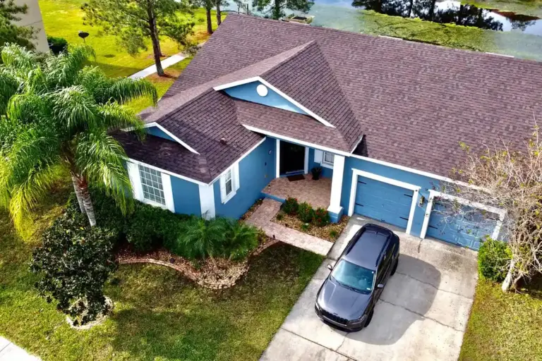 Aerial view of a blue suburban house with a two-car garage and a driveway; a dark car is parked in front, surrounded by green lawn and trees.