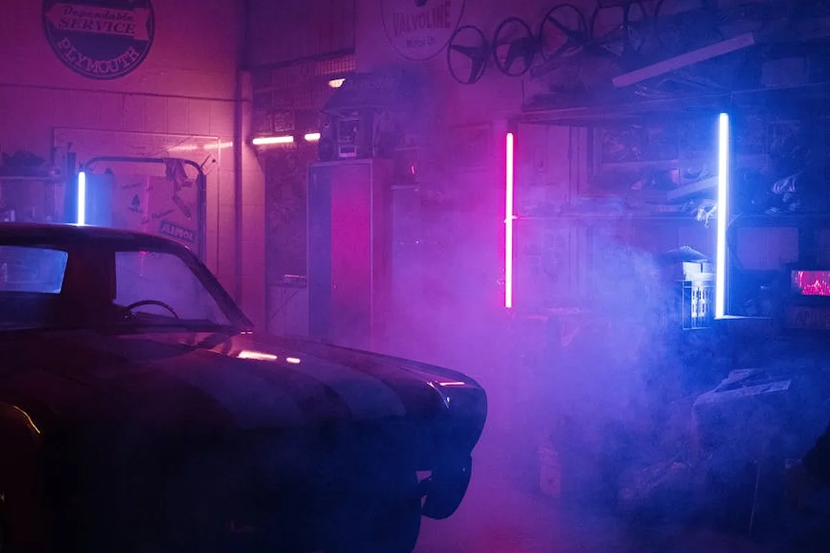 A dimly lit garage with a classic car on the left, neon vertical light tubes in pink and blue casting glow, and a smoky atmosphere.