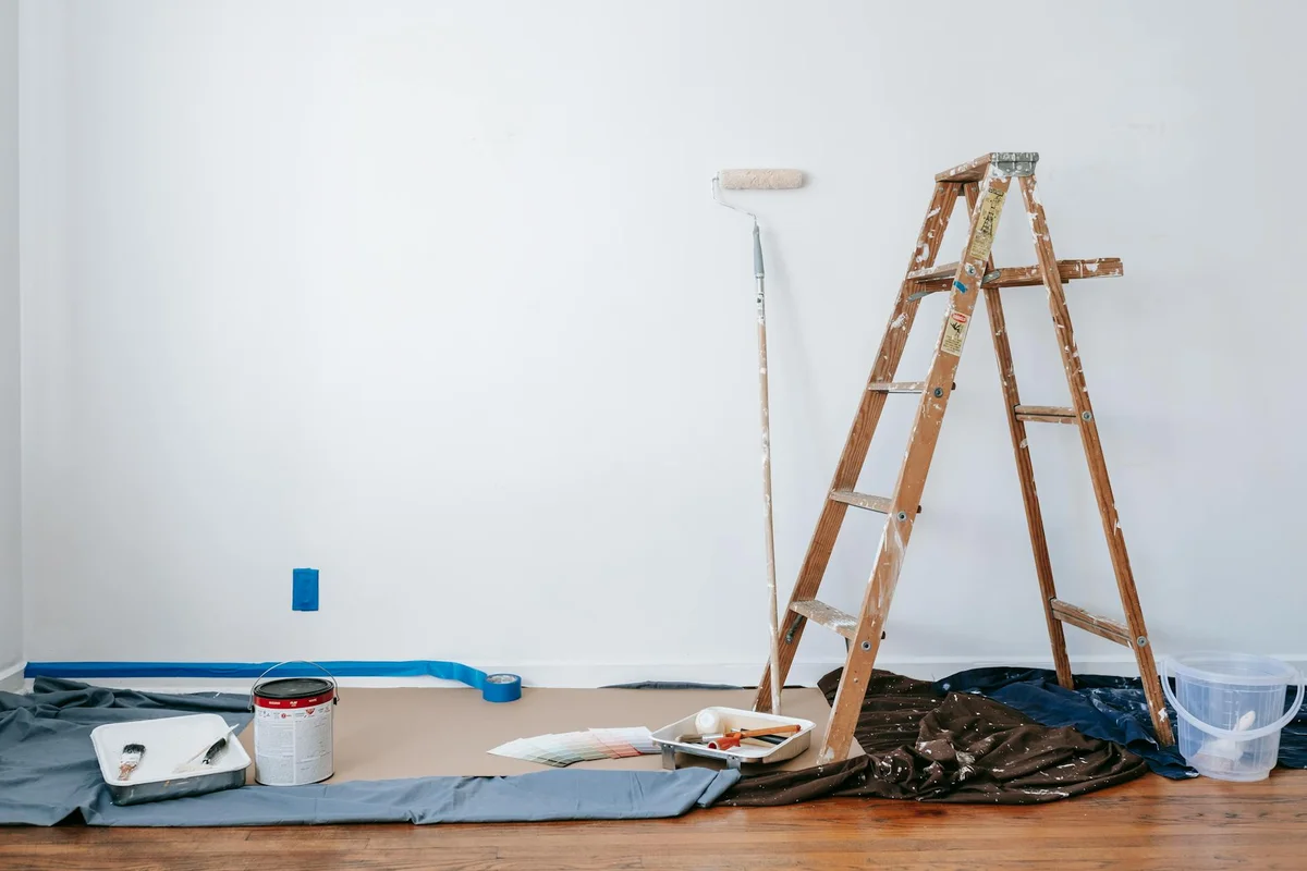 A garage workspace with a wooden ladder, paint roller, a paint tray, buckets, and a drop cloth laid out on the floor, illustrating preparation for diagnosing paint issues.