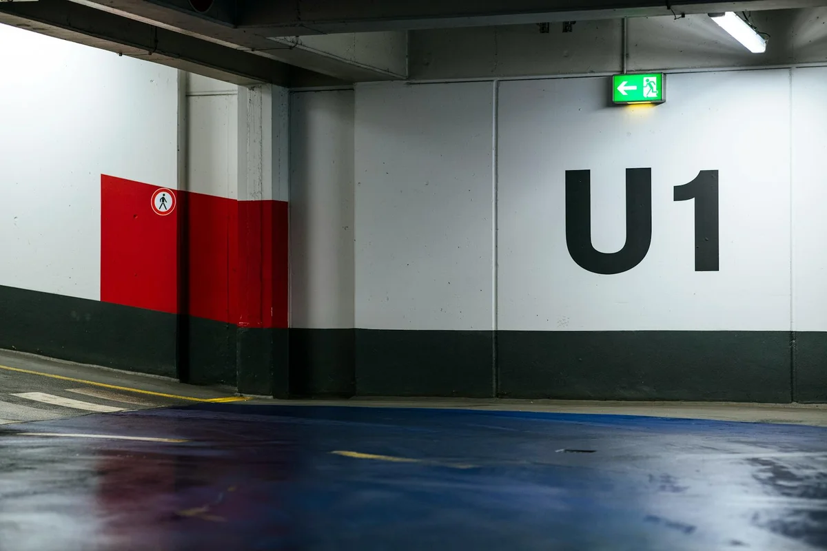 Underground parking garage wall with a bold 'U1' marking, red safety panel on the left, a green exit sign overhead, and a wet, reflective floor.