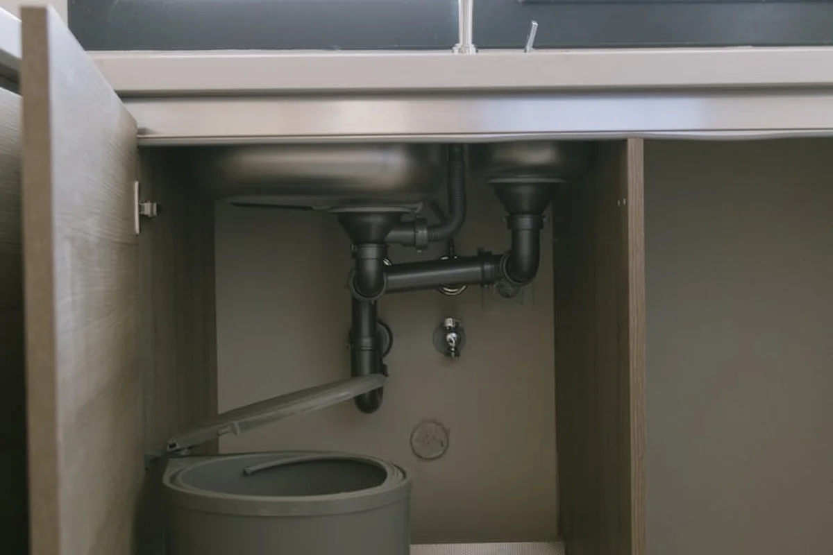 Exposed plumbing pipes under a sink inside a garage cabinet, including a P-trap and drain lines