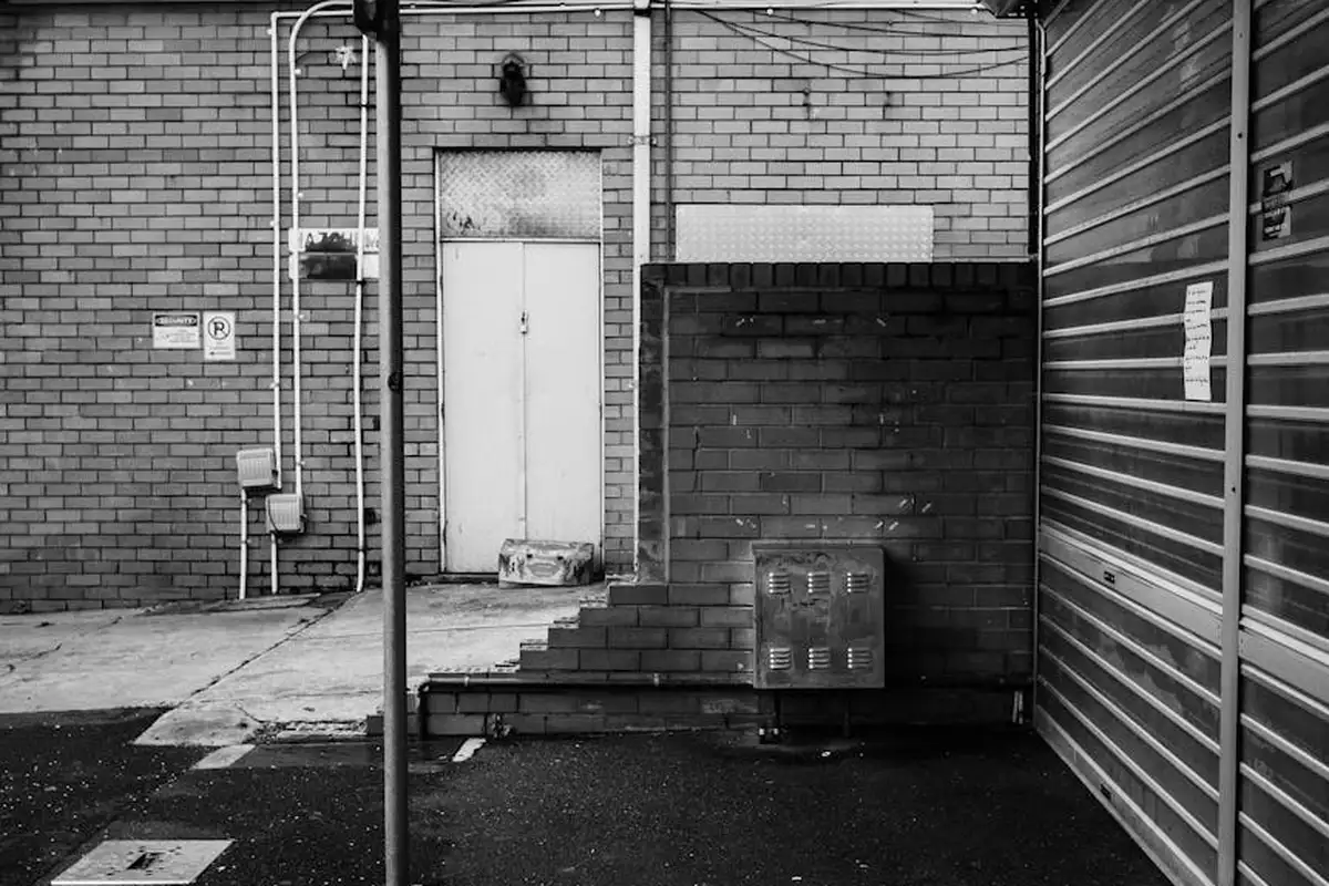 Black-and-white exterior garage area with brick walls, a central door, a metal rolling door on the right, and exposed utility boxes and pipes, suggesting prep space for a smart hub setup.