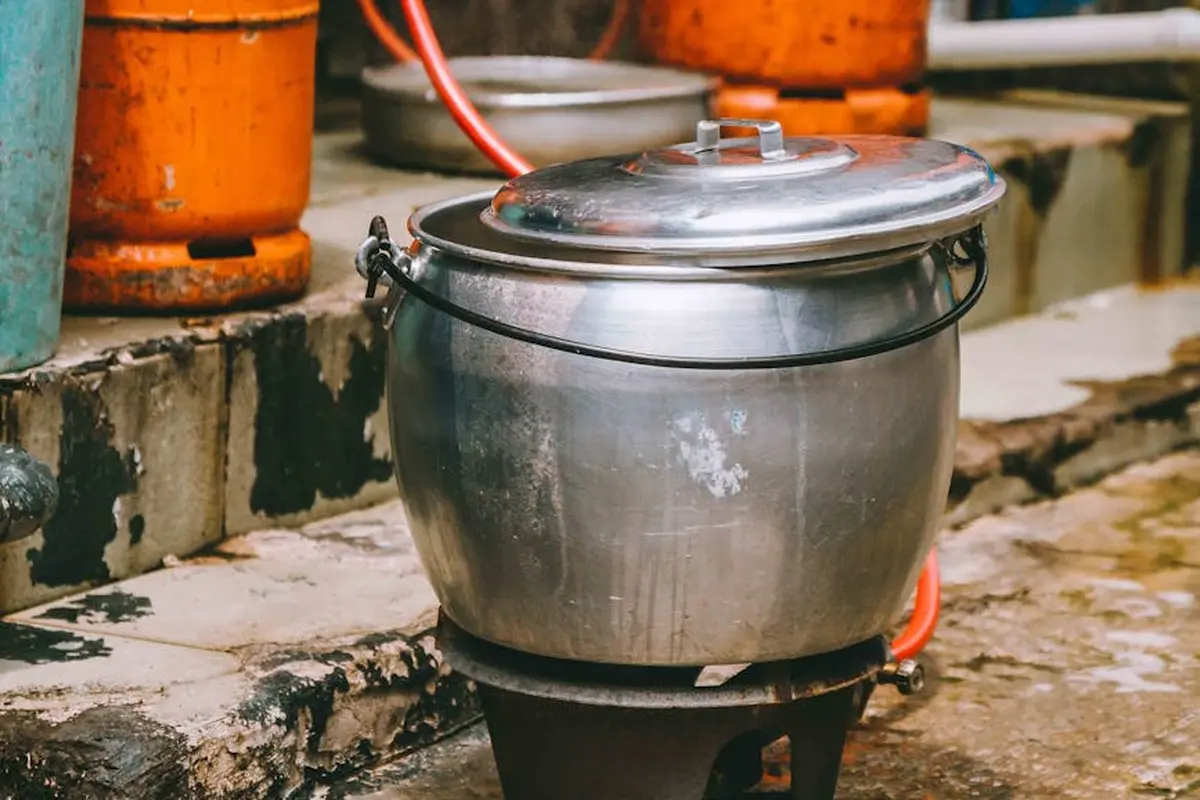 Metal pot with lid on a small portable propane burner outdoors, with orange propane tanks in the background.