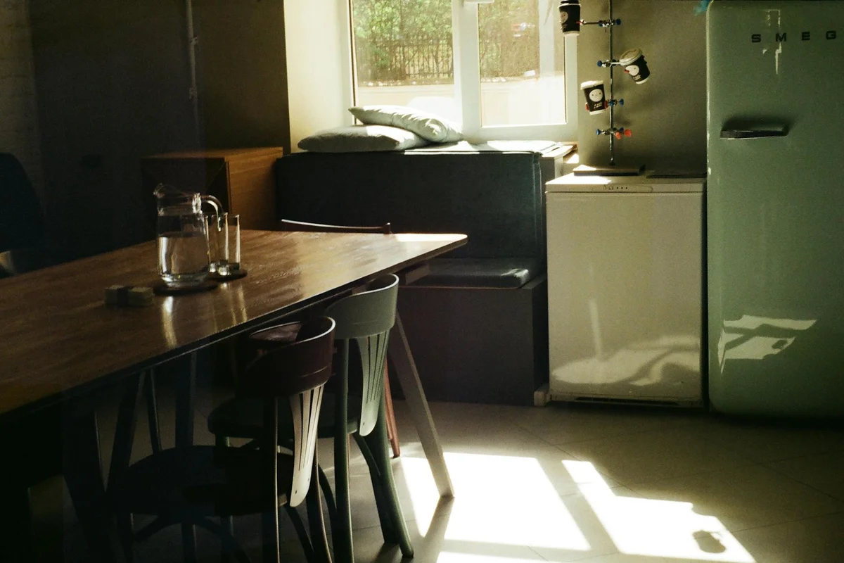 A garage-style room with a tall refrigerator/freezer beside a wooden table and chairs, sunlight streaming through a window.