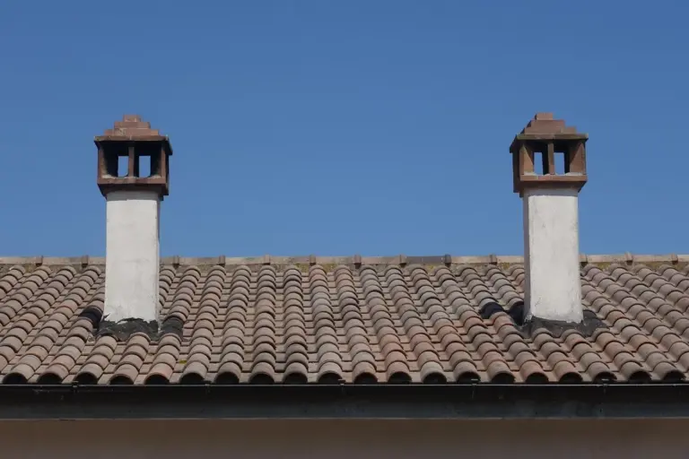 Two chimney flues protrude from a clay-tiled garage roof beneath a clear blue sky.