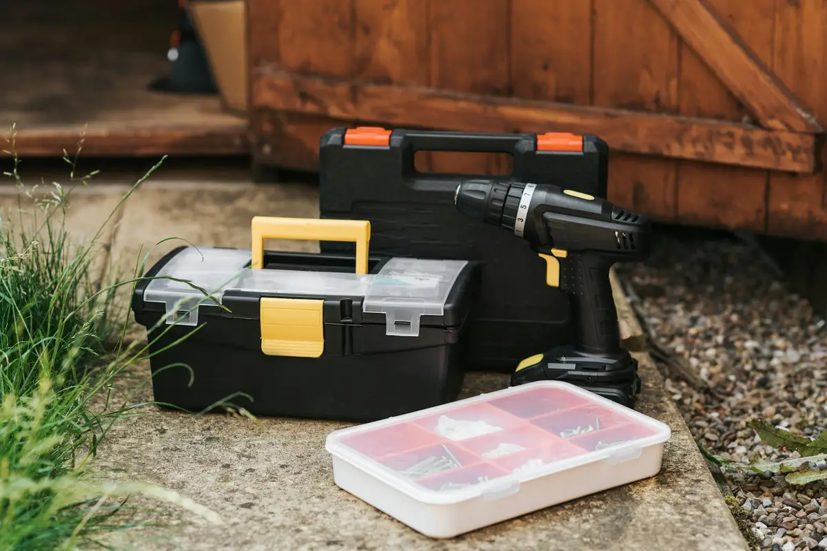 A black toolbox with a yellow latch, a drill, and a clear plastic parts tray sit on a concrete garage floor near a wooden shed door, with some grass nearby.