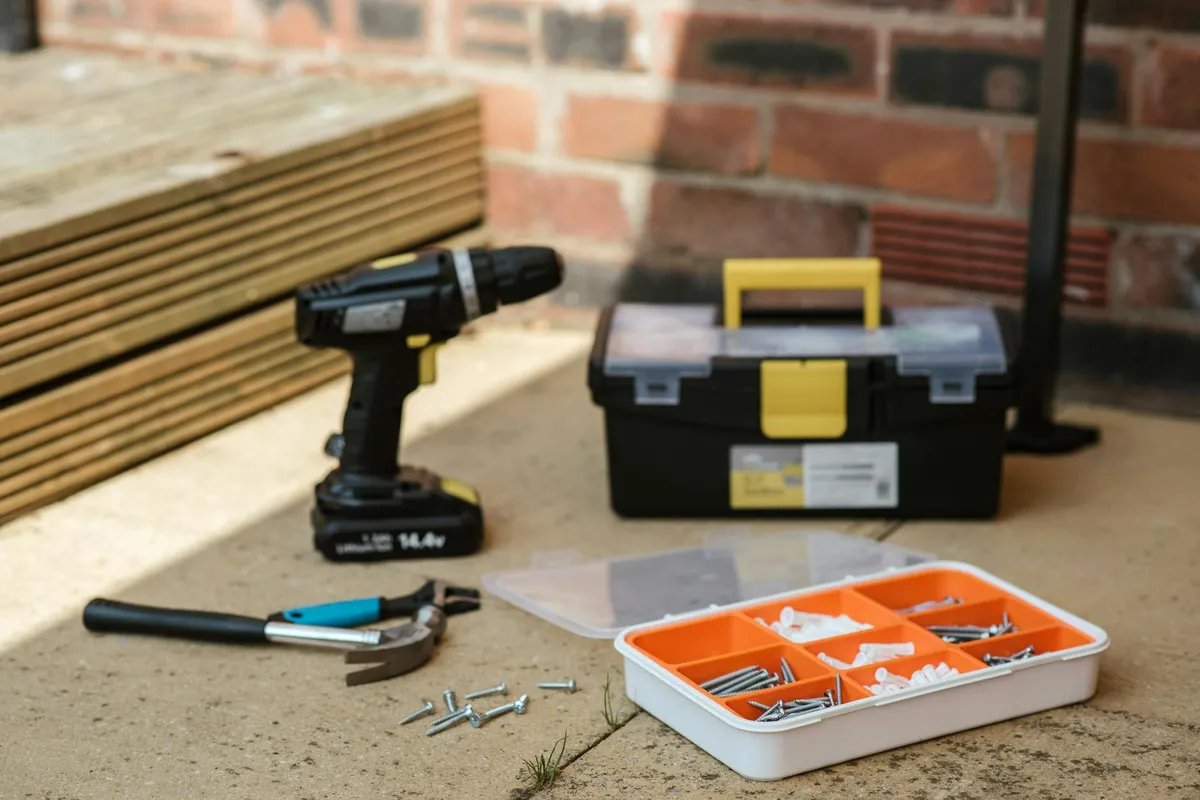 Cordless drill, yellow-and-black toolbox, small organizer with nails and screws, and loose fasteners on a concrete surface beside a brick wall.