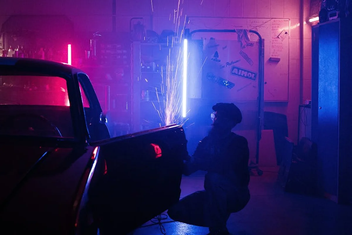 A welder wearing a protective helmet crouches beside a car in a dark garage, illuminated by blue and pink neon lights as sparks fly from welding.