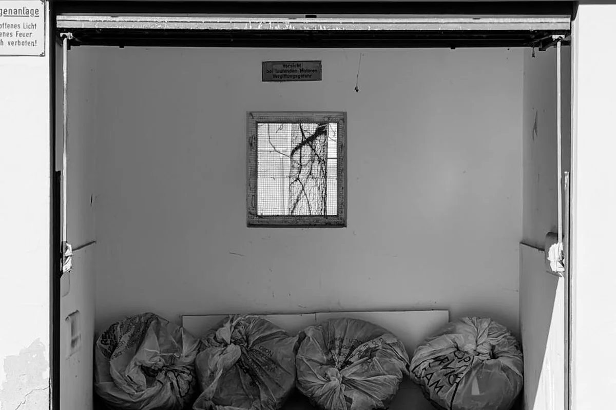 Interior view of a garage with a small square window installed in the wall; bags and clutter sit on the floor beneath the opening.