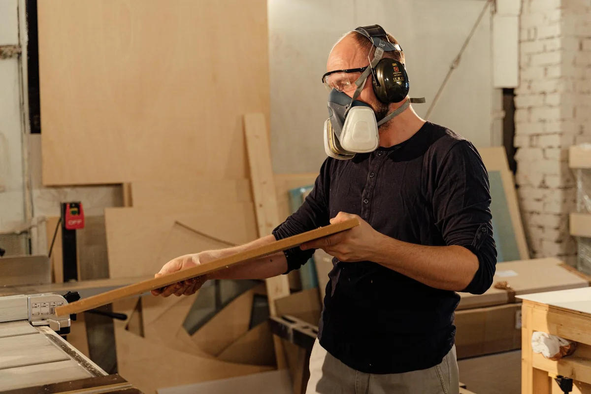 A woodworker wearing a respirator and hearing protection holds a wooden board next to a table saw in a garage-style workshop, with lumber and other tools visible in the background.