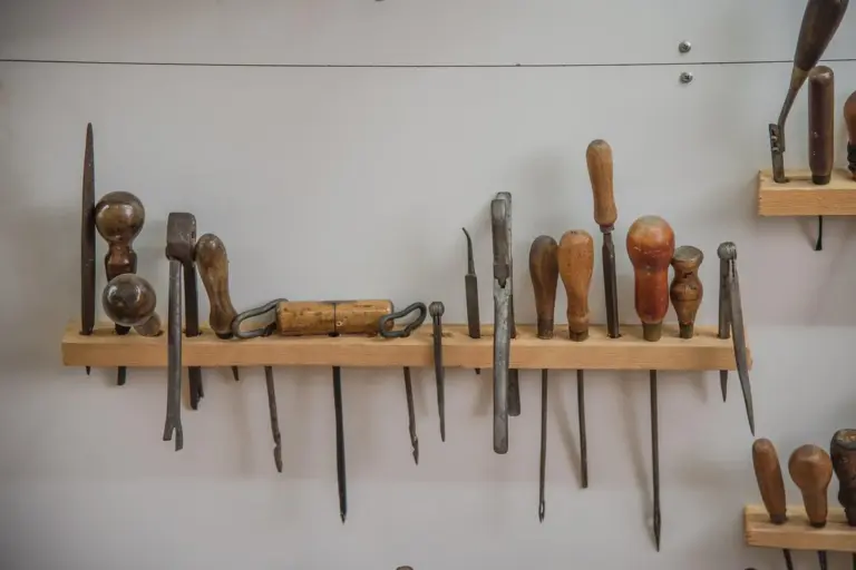 Woodworking chisels, files, and other hand tools hanging on a wooden rail against a gray workshop wall, illustrating organized tool storage.