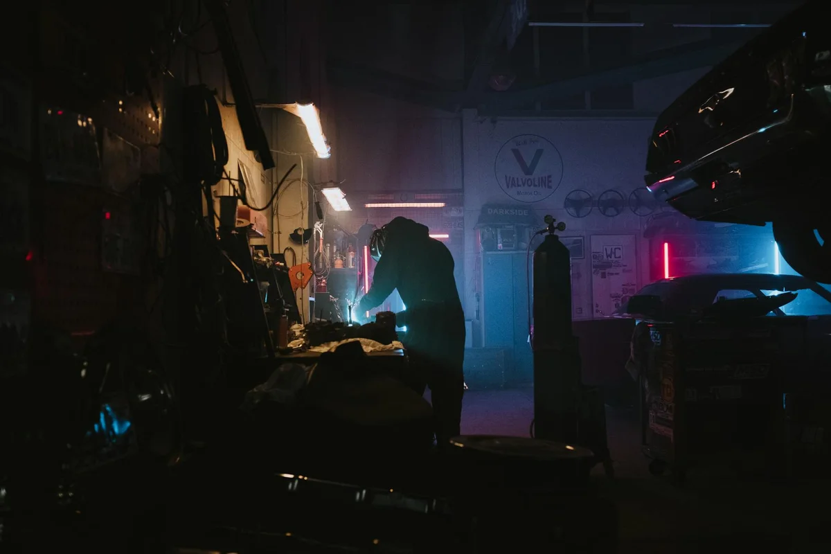 A person works at a cluttered workshop bench in a dimly lit garage, with a car elevated nearby and bright task lights casting blue and orange hues.
