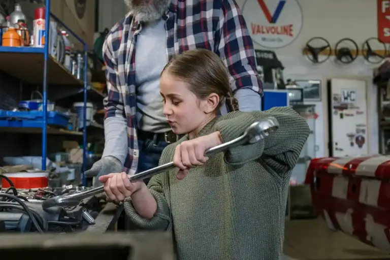 Woman in a green sweater using a wrench on a motorcycle handlebar in a busy garage workshop, with tool shelves and a man in a plaid shirt nearby.