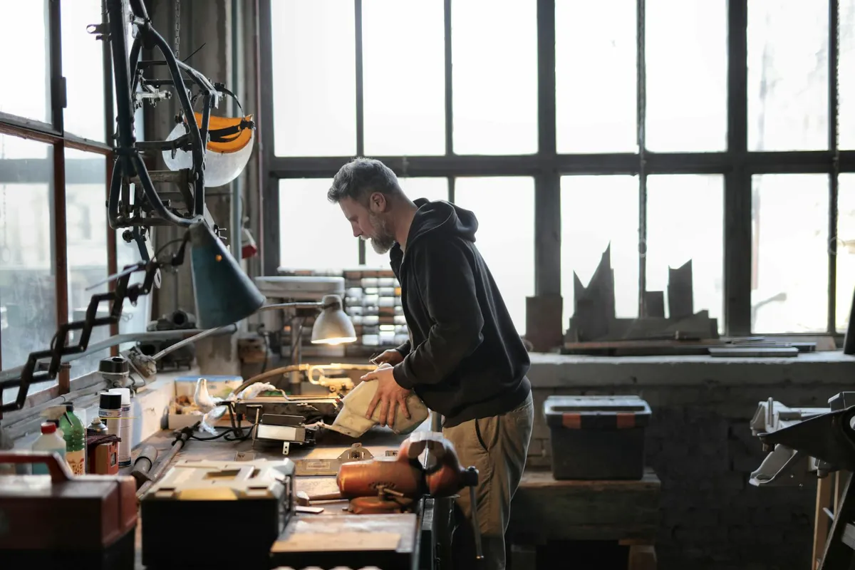 A man in a dark hoodie stands at a cluttered workbench in a bright workshop, examining plans with tools and equipment scattered around.