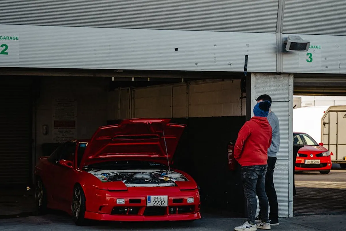 Two people stand in a garage near a red car with its hood open, a second car parked in the doorway, suggesting a hands-on workshop environment.