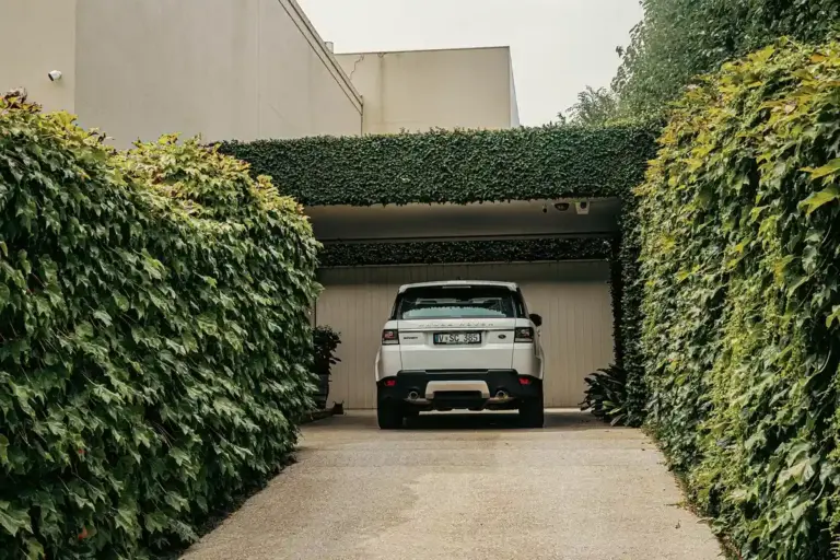 White SUV parked in a driveway facing a closed garage door, with tall green hedges on either side.