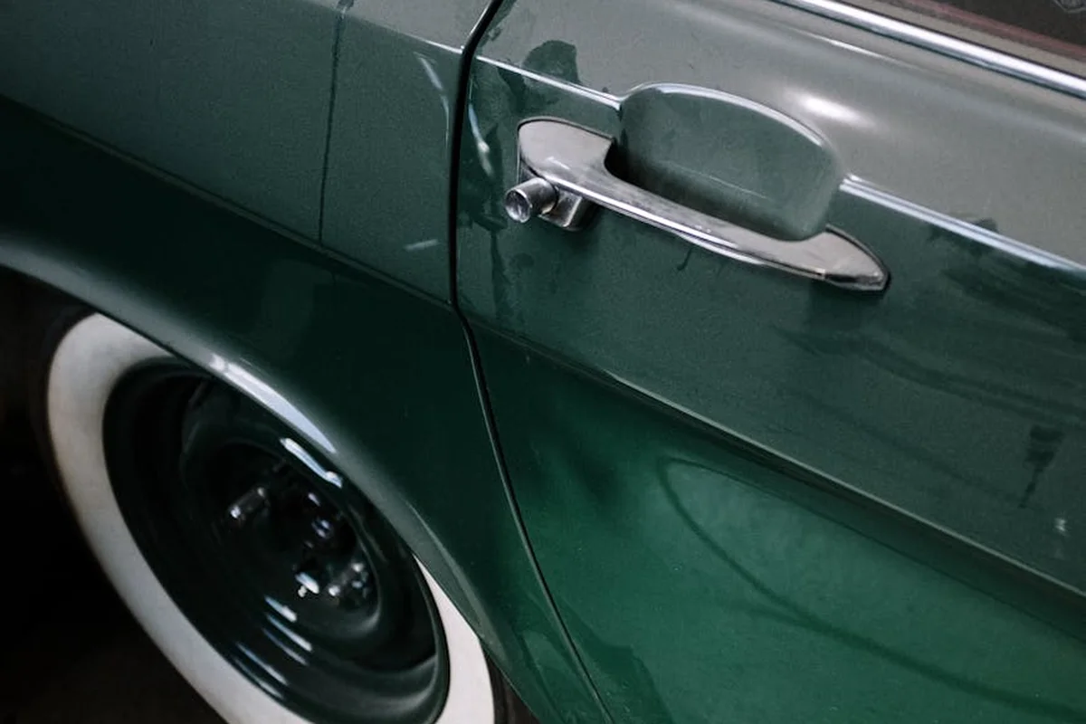 Close-up of a dark green vintage car door with a chrome handle and whitewall tire.