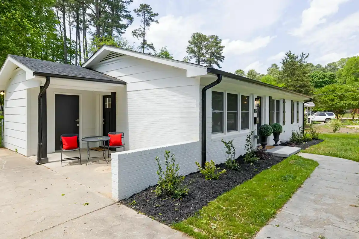 White single-story house with a small porch and red chairs on the patio, a curved driveway, and trees in the background.