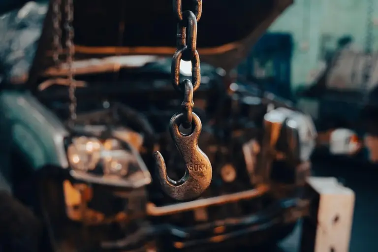 Close-up of a heavy metal hoist hook and chain in a garage workshop with a blurred vehicle in the background.