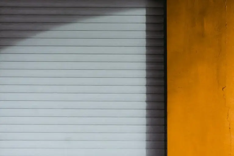 Closed white horizontal roll-up garage door with a yellow wall on the right and a diagonal shadow across the door.