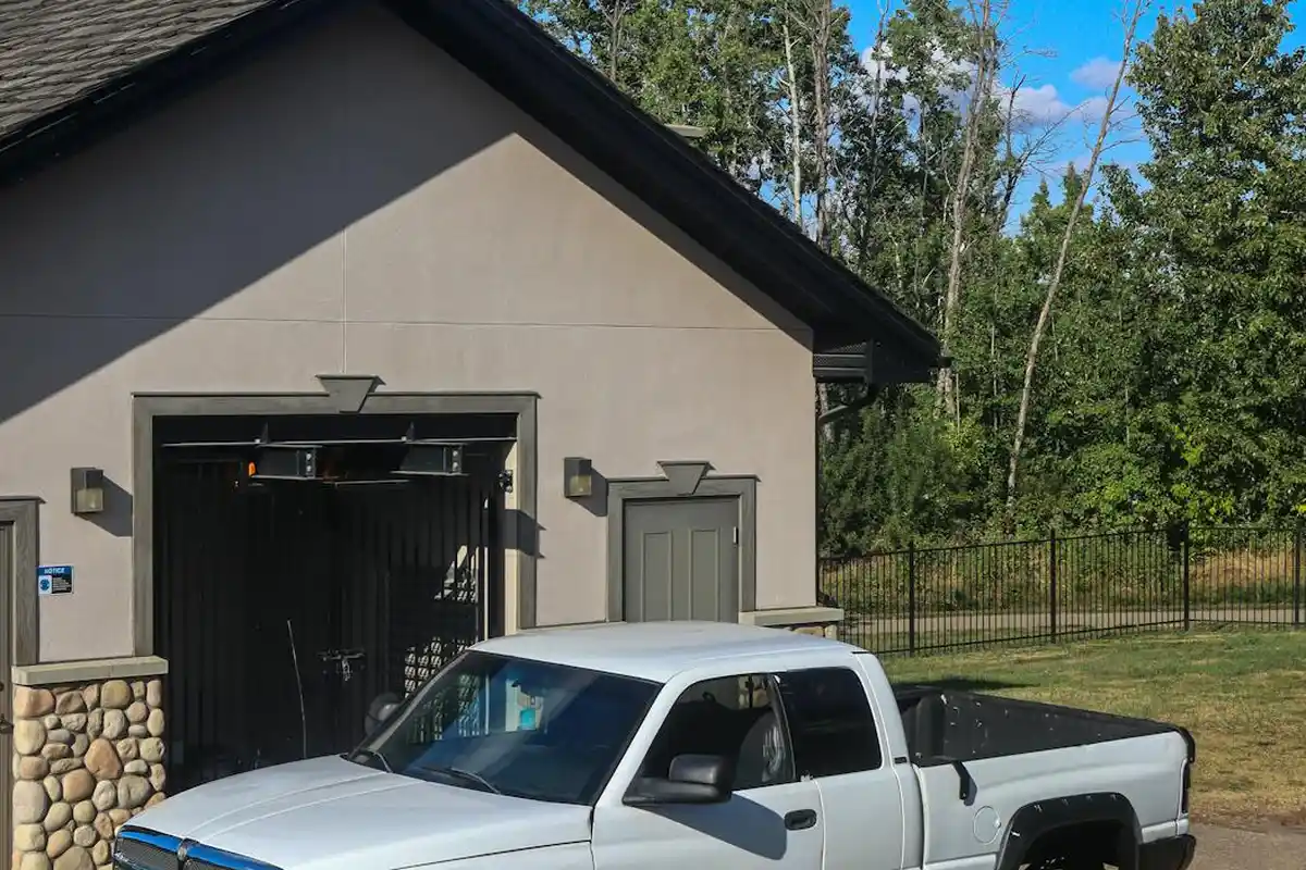 White pickup truck parked in front of a gray residential garage door, daytime with trees in the background