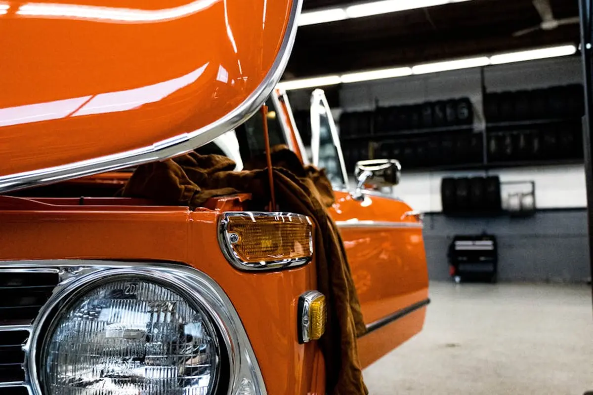 Close-up of an orange vintage car with its hood open inside a garage workshop.